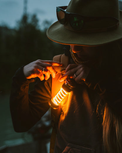 Person adjusts Barebones Edison Light Stick on backpack, its warm glow illuminating the scene, showcasing its dual function as an outdoor lantern and emergency flashlight.
