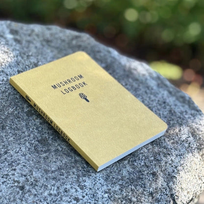 Mushroom Hunter Bundle notebook on rock, titled “MUSHROOM LOGBOOK,” for recording mushroom foraging insights, complements other bundle items like socks and a hunter's knife.