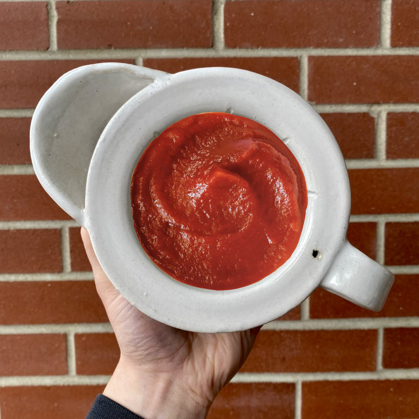 Hand holding a Kent'z Ketchup Shaving Cream 150g ceramic pitcher filled with thick red sauce, emphasizing the product's unique blend of shaving cream and ketchup.