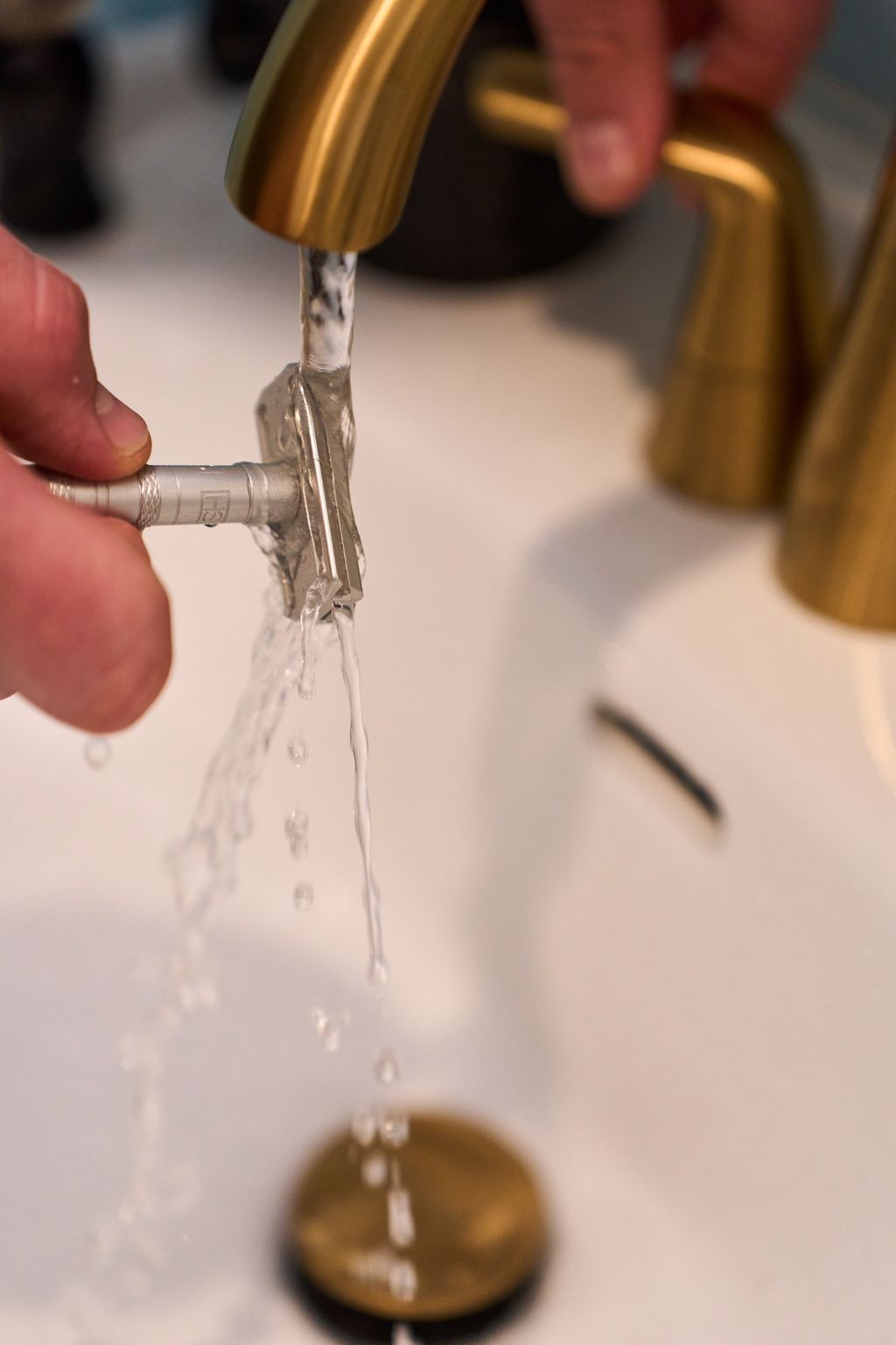 A hand rinses a metal safety razor from Kent’s “Better” Shaving Kit under running water from a gold faucet in a bathroom sink. Water splashes off the razor as another hand adjusts the tap handle, highlighting the elegance of Kent’s “Better” Shaving Kit in use.