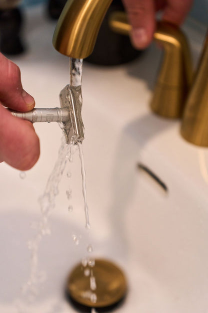 A hand rinses a metal safety razor from Kent’s “Better” Shaving Kit under running water from a gold faucet in a bathroom sink. Water splashes off the razor as another hand adjusts the tap handle, highlighting the elegance of Kent’s “Better” Shaving Kit in use.
