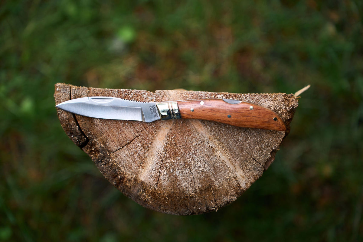The Grohmann Mini Russell Lockblade with Rosewood Handle, featuring a 4″ clip point stainless steel blade, rests atop a cut tree stump amidst green grass. Its blade glints in the sunlight, embodying the renowned craftsmanship of Grohmann knives.