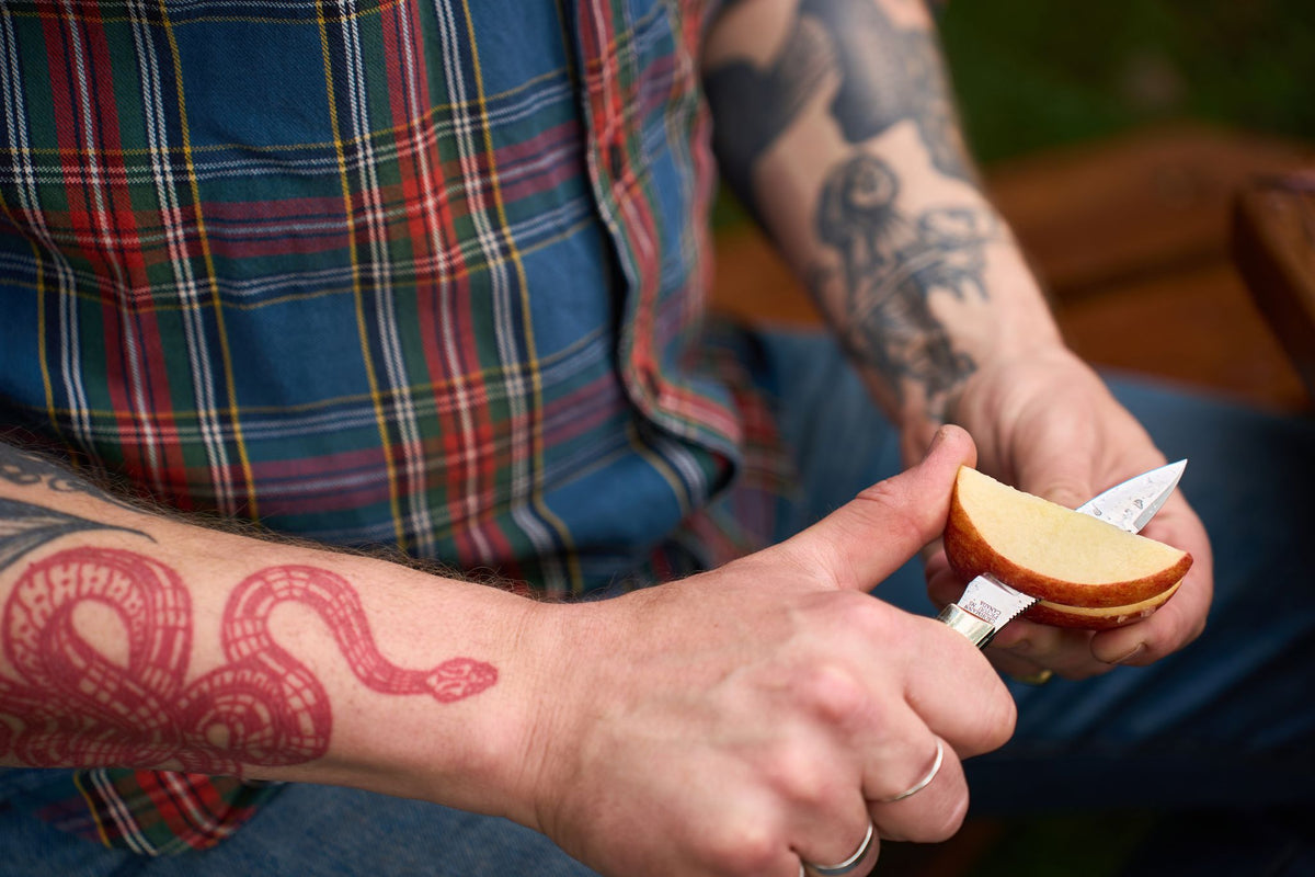 A person with tattoos, dressed in a plaid shirt and jeans, sits outside slicing an apple with the Grohmann Mini Russell Lockblade. The 4″ clip point stainless steel blade and rosewood handle let them make precise cuts.