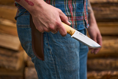 A person in jeans and a plaid shirt holds a Helle Knives Nord with a wooden handle, standing near stacked logs—a scene that embodies Scandinavian style and the spirit of survival and bushcraft.