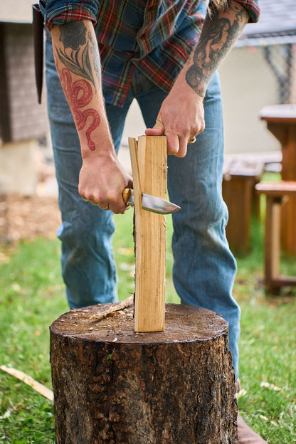 A person with tattooed arms splits a piece of wood using the Helle Knives Nord, pressing it onto a tree stump outdoors. Dressed in jeans and a plaid shirt, they demonstrate essential survival and bushcraft skills in true Scandinavian style.