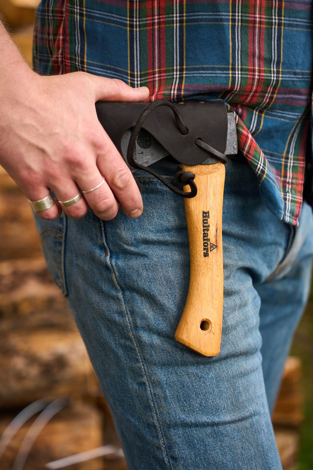 A person wearing jeans and a plaid shirt holds a small Hultafors Hultan Hatchet with a wooden handle and sheath, tucked into their back pocket.
