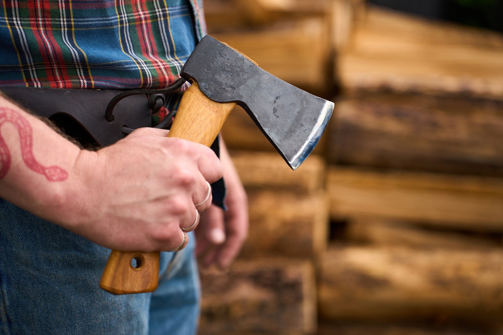 A person wearing jeans and a plaid shirt holds a Hultafors Hultan Hatchet with a wooden handle in front of stacked firewood.