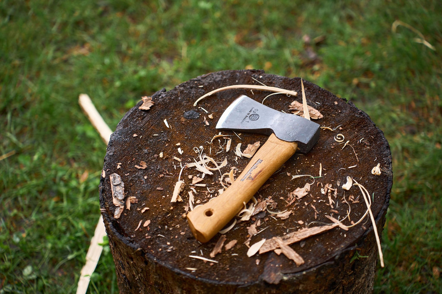 A Hultafors Hultan Hatchet with a sturdy wooden handle rests on a tree stump, surrounded by wood shavings, with green grass in the background.