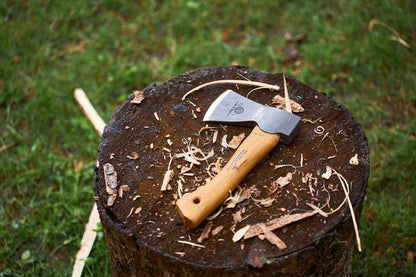 A Hultafors Hultan Hatchet with a sturdy wooden handle rests on a tree stump, surrounded by wood shavings, with green grass in the background.