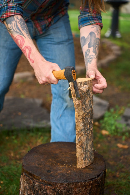 A person with tattoos uses a Hultafors Hultan Hatchet to split a log on a tree stump outdoors. Only their arms and torso are visible; they’re wearing jeans and a plaid shirt.