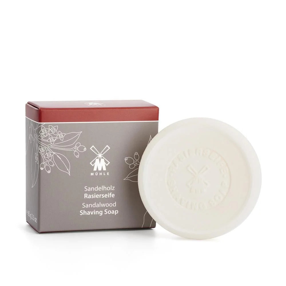 A round, white Mühle Sandalwood Shaving Soap bar is displayed in front of its gray box, which features botanical illustrations and white text. The soap is vegan and has an embossed logo.