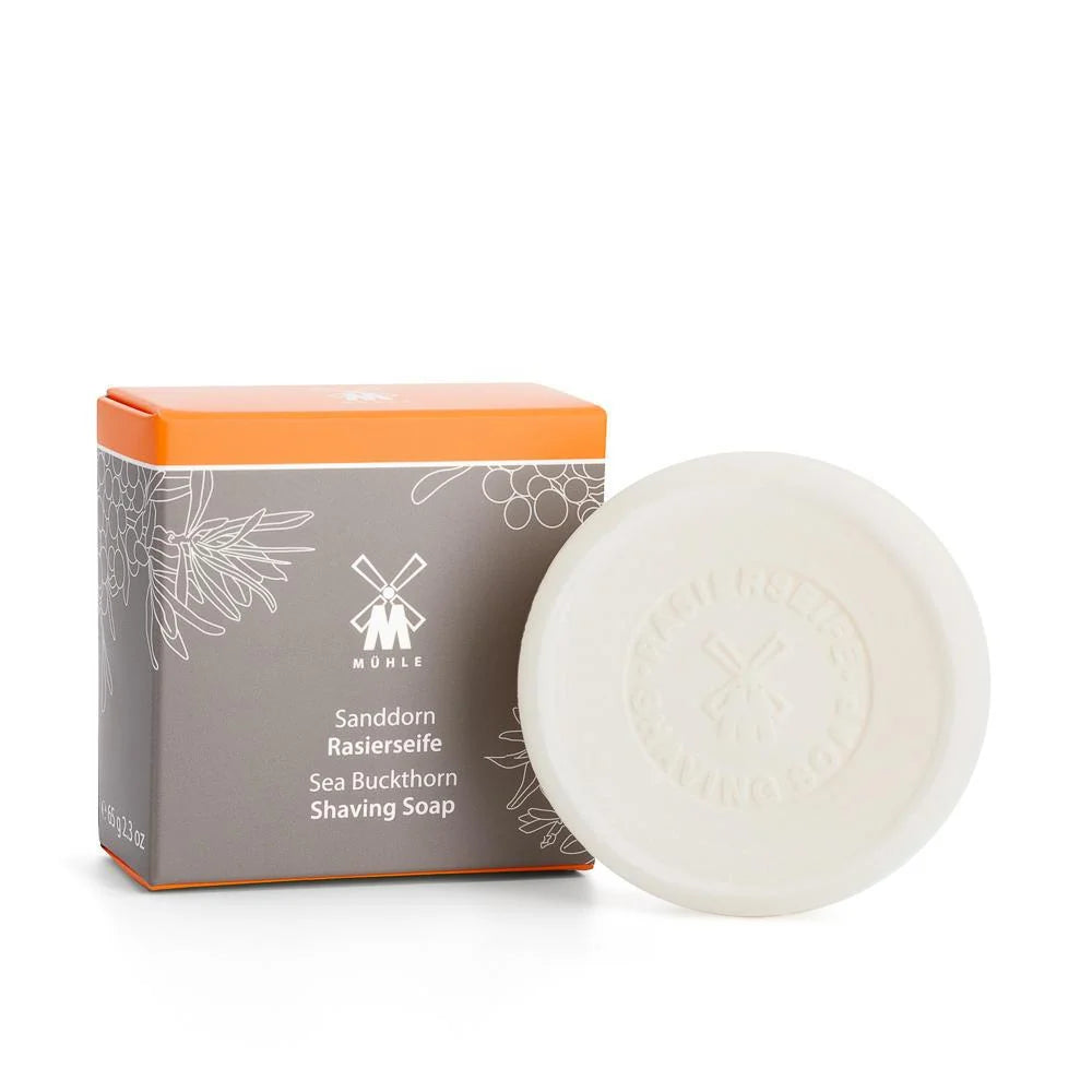 A round bar of Mühle Sea Buckthorn Shaving Soap is displayed in front of its box, which is gray with orange accents and adorned with a botanical illustration. The packaging highlights the product’s plastic-free design and features the Sea Buckthorn Shaving Soap label.