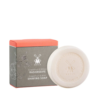 A round white shaving soap featuring embossed text sits in front of a gray and orange box decorated with floral and fruit motifs. The packaging displays “Mühle Grapefruit & Mint Shaving Soap” in both English and German, emphasizing this vegan soap’s luxurious and refreshing grapefruit mint scent.