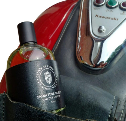 Crown Shaving Co. Eau de Parfum 100mL tucked in a black leather pouch, with a red Kawasaki motorcycle seat and chrome console partially visible in the background.
