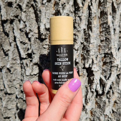 Hand with lavender nails holds Boreal Folk Tallow Skin Stick, a natural skin balm, against tree bark, highlighting its eco-friendly packaging and Canadian-sourced ingredients.