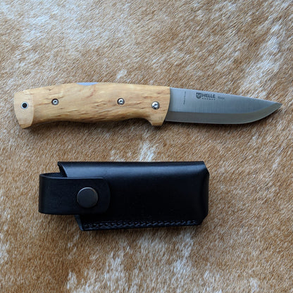 Helle Knives Bleja 85mm Folding Knife with light wood handle on animal-hide surface, accompanied by its black leather sheath.