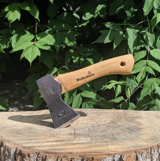 Hultafors Agelsjon Mini Hatchet with a wooden handle embedded in a tree-stump, ideal for camping and hiking tasks, showcased against leafy foliage.
