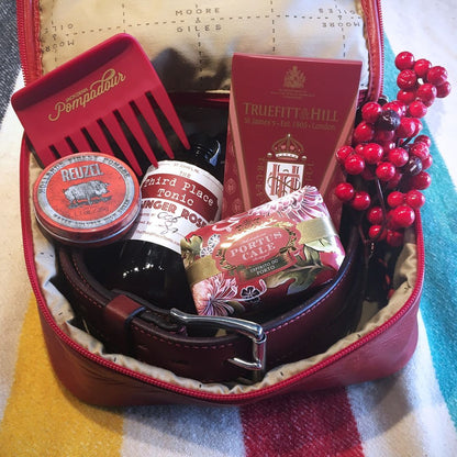 Reuzel Pomade Regular Red tin in a leather travel kit with grooming essentials, including a comb, hair tonic, soap, and shaving product, on a wool blanket.
