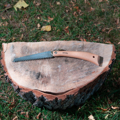 Opinel Folding Saw with wooden handle and serrated stainless steel blade, open on a tree stump surrounded by grass and leaves, ideal for outdoor tasks.