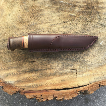 Helle Knives Harding 100mm Hunting Knife sheathed in handcrafted leather, displayed on a rustic wood slab, showcasing its dual-tone wooden handle with decorative spacers.