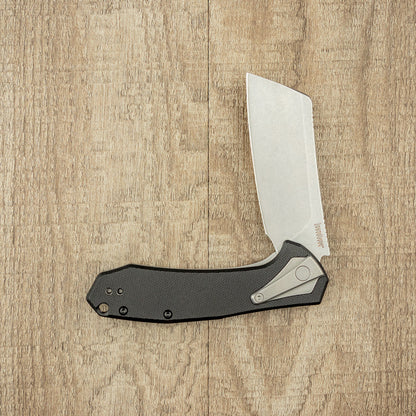 Kershaw Bracket folding mini-cleaver with a broad, stone-washed blade and black textured handle, featuring a metal pocket clip, lying open on wooden surface.