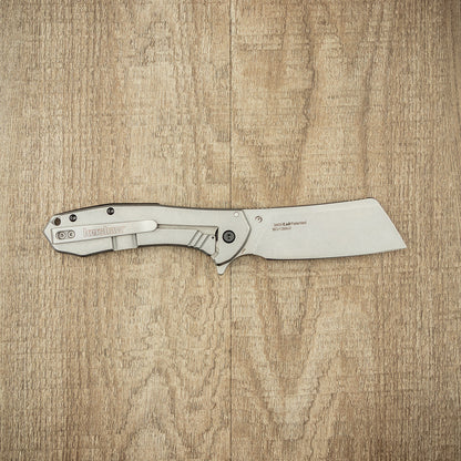 Kershaw Bracket pocket-knife on a wooden surface, featuring a cleaver-style drop-point blade and metal handle with pocket clip, ideal for versatile daily and campfire use.
