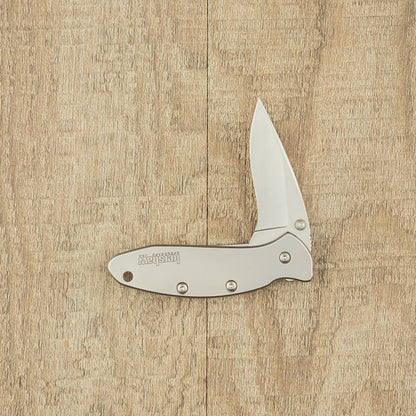Kershaw Chive pocket knife open on wood surface, silver metal design with Kershaw on handle, ideal for small tasks and easy portability.