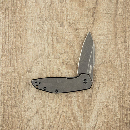 Kershaw Gravel folding pocket knife with a stonewashed-finish blade and black handle, showcased on a wood-grain surface, highlighting its durability and industrial design.