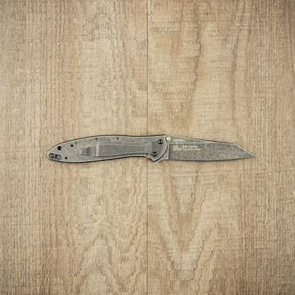 Kershaw Random Leek Blackwash folding pocket knife, open on wooden surface, showcasing weathered metal handle and blade, designed by Ken Onion for precise, reliable use.