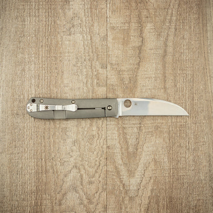 Spyderco Swayback Pocket Knife with a titanium handle, round thumb-hole for one-handed opening, and slim, straight-edged blade, displayed on a wooden surface.