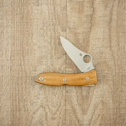 Spyderco SpyOpera Pocket Knife, featuring a satin-finished drop-point blade and tan Micarta handle, displayed partially open on a wood-grain surface, showcasing its sleek design.