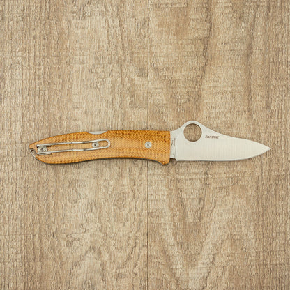 Spyderco SpyOpera Pocket Knife with a micarta handle, M390 steel blade stamped “LionSTEEL,” and wire pocket clip, displayed on a wood-grain surface.