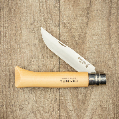 Opinel Inox No.10 Folding Knife with stainless-steel blade and wooden handle, open on wooden surface, exemplifies timeless design and reliable craftsmanship from a renowned French brand.