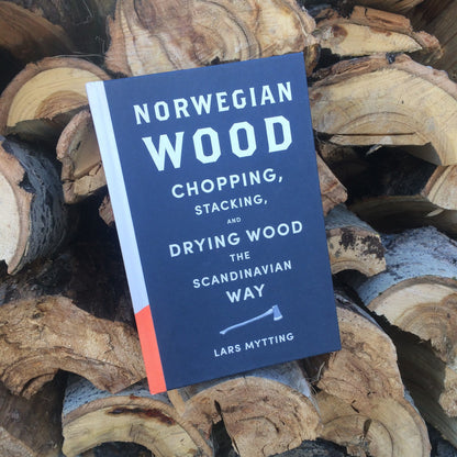 Hardcover book titled Norwegian Wood rests on stacked firewood, part of the Ultimate Axe Bundle, featuring essential tools and guides for wood chopping enthusiasts.