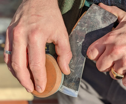 Close-up of hands honing a metal tool with a circular stone, illustrating techniques taught in Kent of Inglewood's Axe & Hatchet Sharpening Classes.