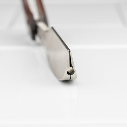 Feather Artist Club DX Folding Handle Shaving Razor, showcasing its blade heel and hinge pin, nestled in wooden scales on a glossy surface.