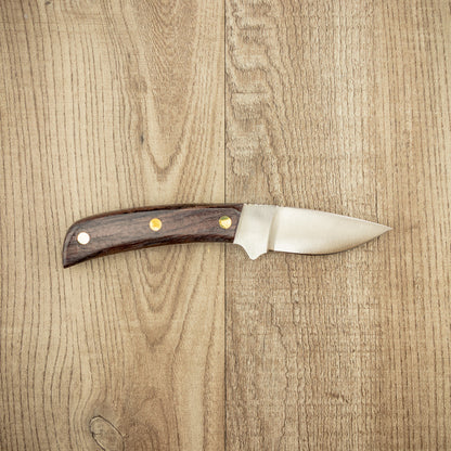 Grohmann #104F Flat Grind Mini Skinner with polished stainless steel blade and rosewood handle, featuring brass rivets, displayed on light wood surface, perfect for controlled animal dressing.