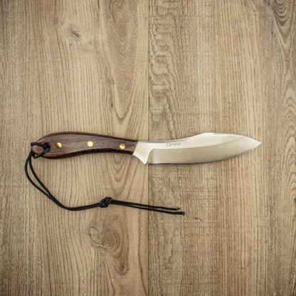 Grohmann #4 Survival Fixed Blade on wood surface, featuring a full-tang stainless steel blade with Canada etched, dark rosewood handle, brass rivets, and black cord lanyard.