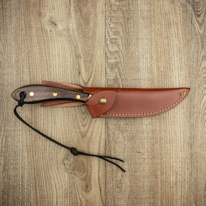 Grohmann #4 Survival Fixed Blade knife with a rosewood handle, brass pins, and lanyard, in a brown leather sheath on a wood-grain surface.