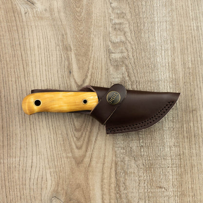 Helle Knives Mandra 70mm Neck Knife with light-wood handle and dark-brown leather sheath, designed by Les Stroud, laid on wood surface.