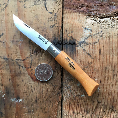 Opinel Carbon No.05 Folding Knife with open carbon steel blade, light wooden handle, and Canadian quarter for scale, displayed on a rustic wooden surface.