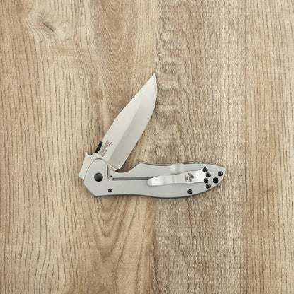 Kershaw Emerson CQC-6K D2 partially opened on a wood surface, showcasing its stainless-steel blade, metal handle, and pocket clip, reflecting Kershaw's quality craftsmanship.
