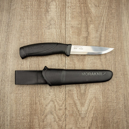Mora Companion 4 Camping Knife with stainless-steel blade and black handle, accompanied by its black sheath, displayed on a wooden surface.