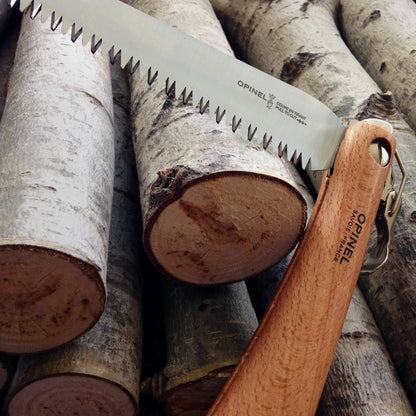 Opinel Folding Saw with a wooden handle, laid on freshly cut birch logs. Ideal for trail clearing and yard work, featuring a stainless steel blade.