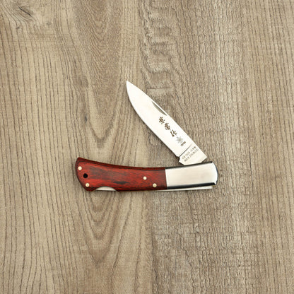 Seki Kanetsune Kaico-Tou Classic Folding Knife 70mm with polished AUS-8 stainless blade and Pakka wood handle, lying open on a wood-grain surface.