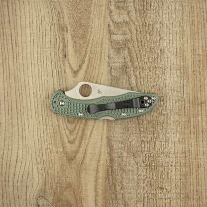 Spyderco Delica 4 folding knife with green FRN handle, black pocket clip, and partially visible VG10 blade with thumb hole, on a wood-grain surface.
