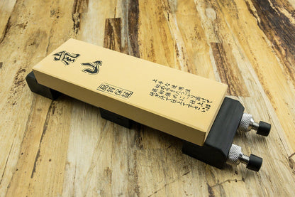 Stone Holder with adjustable plastic jaws and thumbscrews, securing a Japanese-style whetstone on a wooden tabletop, ideal for stable knife or tool sharpening.